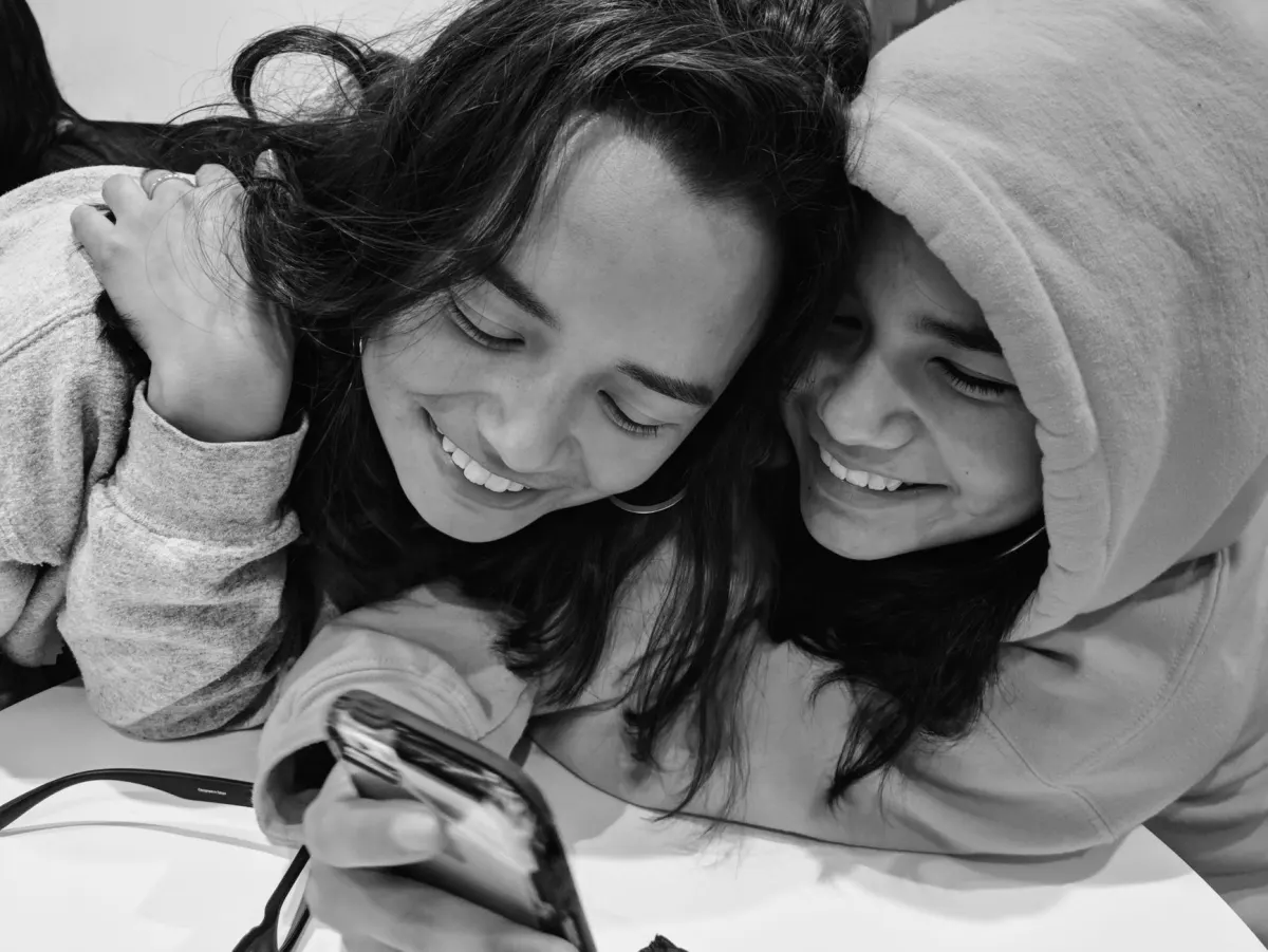 Dois amigos rindo juntos enquanto olham a tela de um celular, refletindo a proximidade que um coração amarelo transmite.