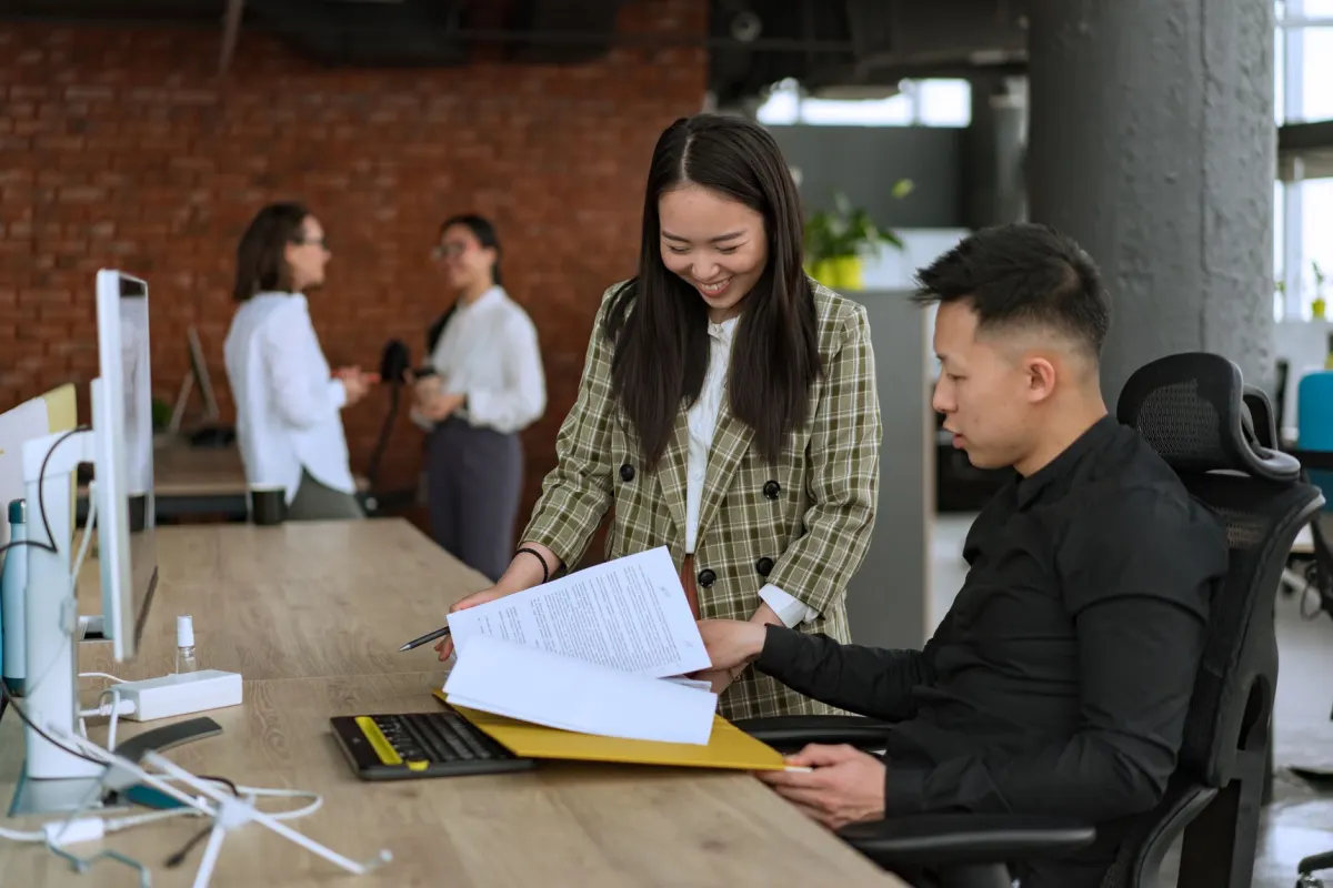 Two coworkers reading a document together at a desk, like a second reviewer checking a draft before a person publishes it.