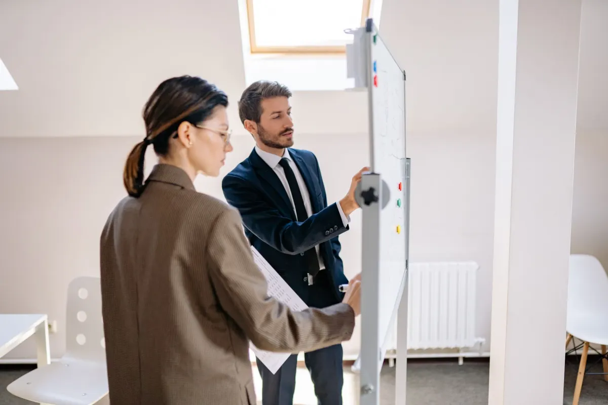 Two people plan a workflow on a whiteboard, reflecting the cost tradeoffs of scheduling and rescheduling a video.