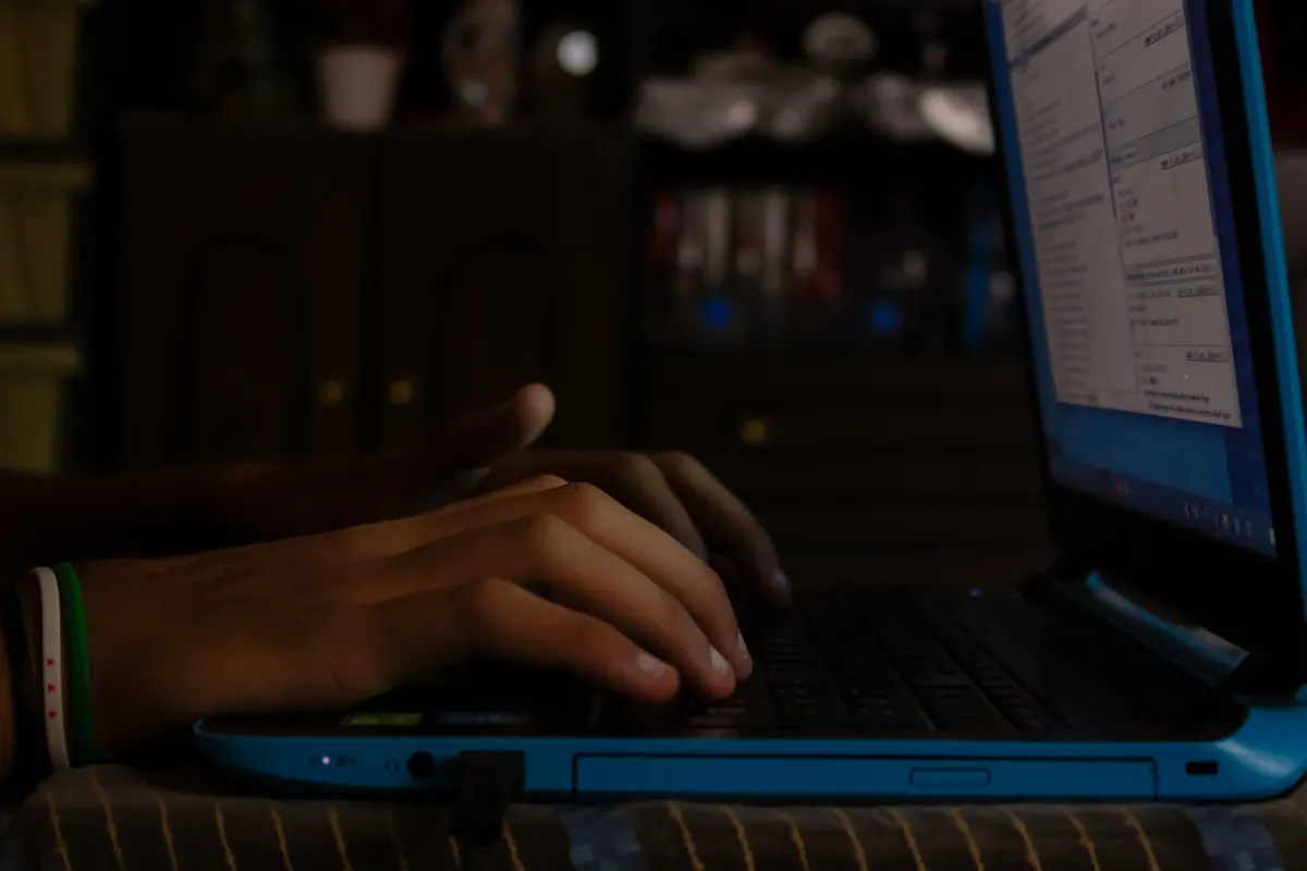 A person typing on a laptop in a dimly lit room, illustrating posting anonymously in a Facebook group.