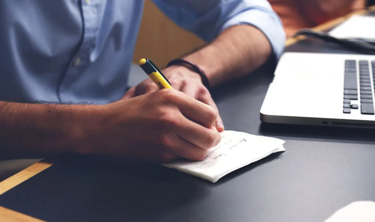 A person writes at a laptop with a notebook nearby, representing the long-term upkeep an evergreen answer needs compared with a single Reddit thread.